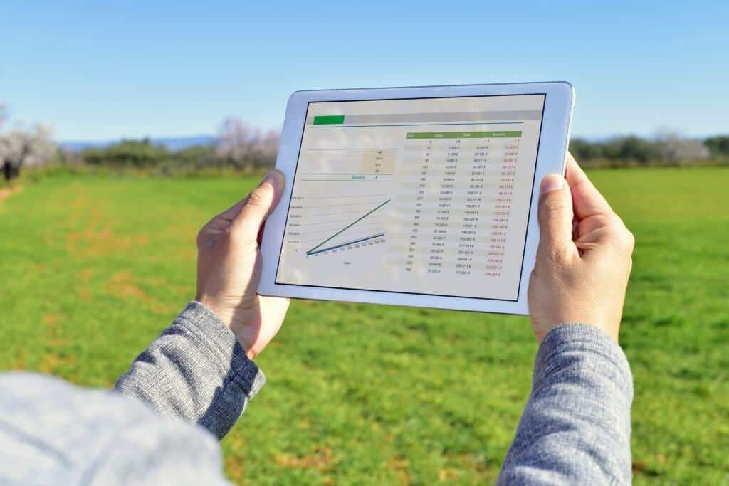 foto no campo que mostra os braços de uma pessoa com moletom cinza segurando um tablet na horizontal que mostra na tela uma planilha de gestão agrícola
