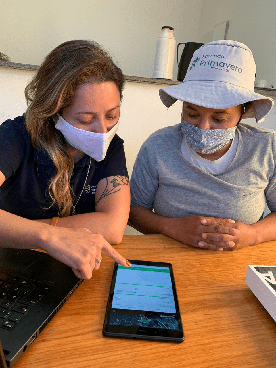 foto de duas mulheres usando máscara de pano, camisetas, sentadas em um escritório fazendo monitoramento de pragas de café e planejamento das operações com insumos pelo aplicativo Aegro. O tablet está numa mesa de madeira.