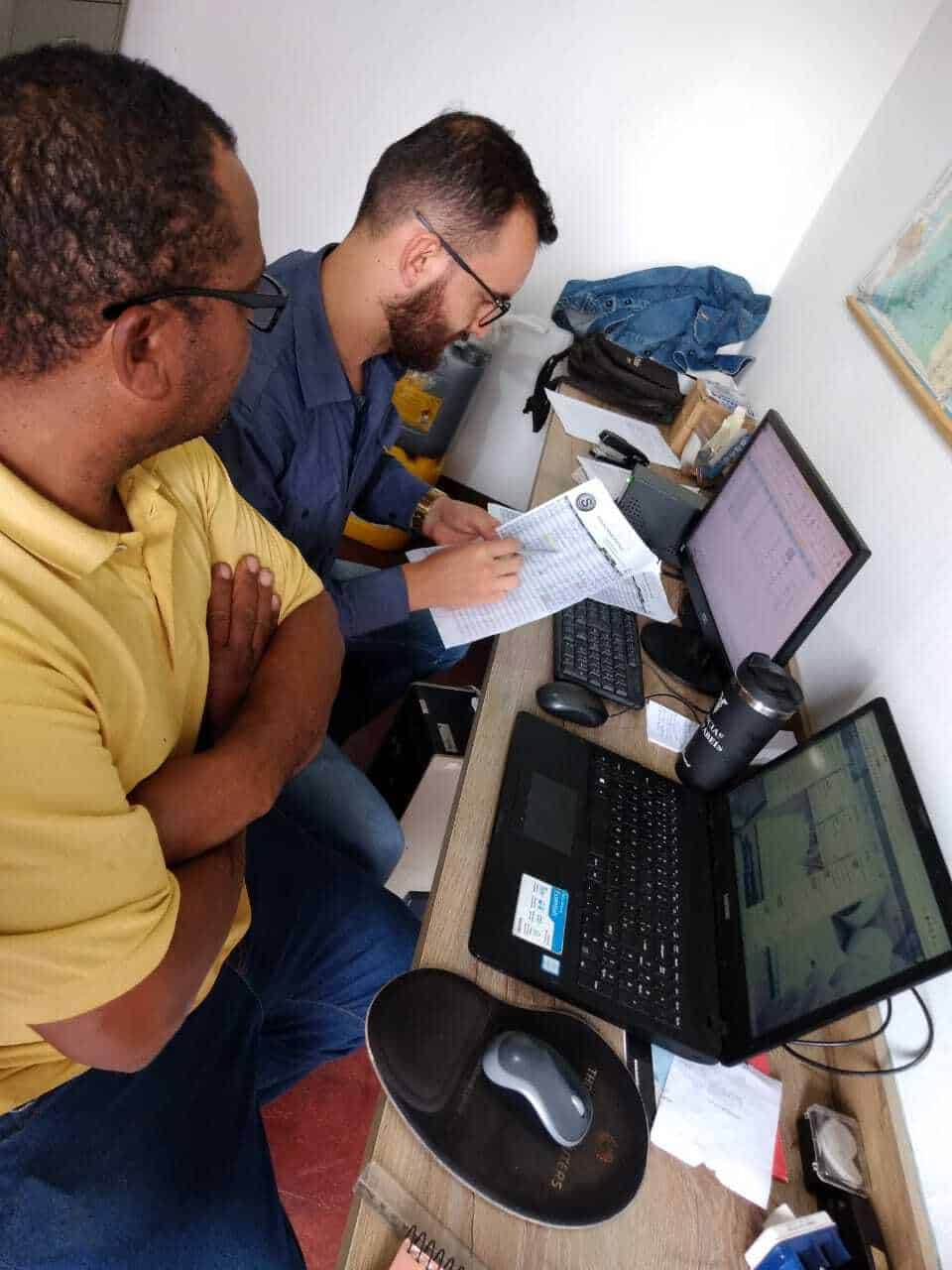 Imagem mostra consultor de controladoria e gestão financeira e funcionário sentados à mesa, em frente a dois computadores. Os dois olham para uma folha de papel.