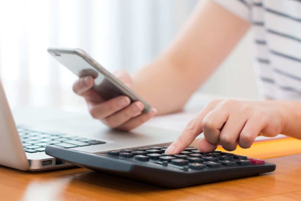 elaboração de projetos de crédito rural: imagem dos braços de uma mulher que mexe no celular e faz cálculos usando uma calculadora. Na frente dela, há um laptop.