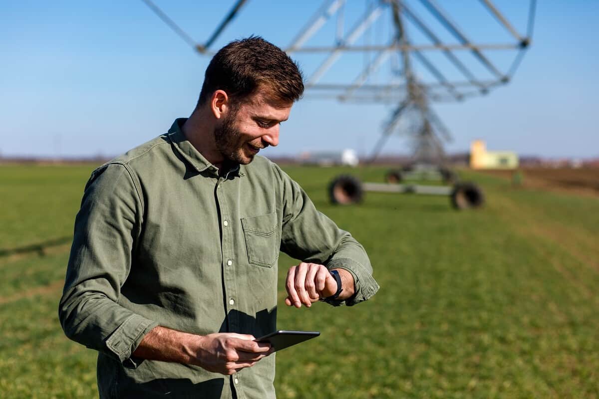 Imagem mostra homem jovem em uma fazenda, sorrindo e olhando para um relógio no pulso.
