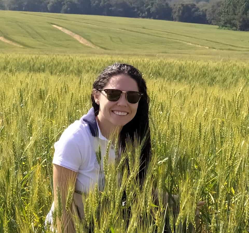 Foto da redatora Carina no meio de uma lavoura de trigo. Ela está com blusa branca, óculos escuros e sorrindo.