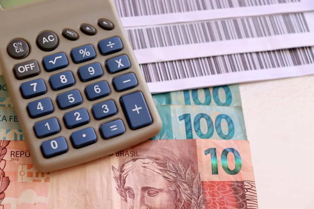 Imagem de calculadora, cédulas de 10 e 100 reais e alguns boletos no fundo