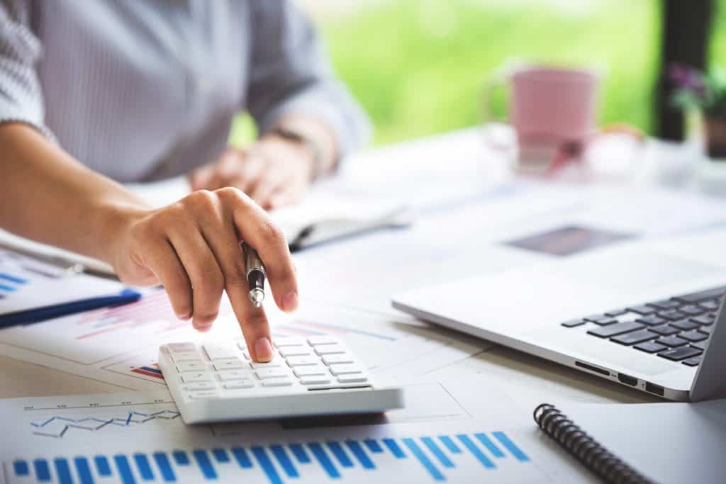 indicadores de produção no Aegro: imagem de mulher digitando em uma calculadora, em uma mesa com fazenda no fundo. Na mesa, há computador, caneca, papéis com gráficos e uma caneta