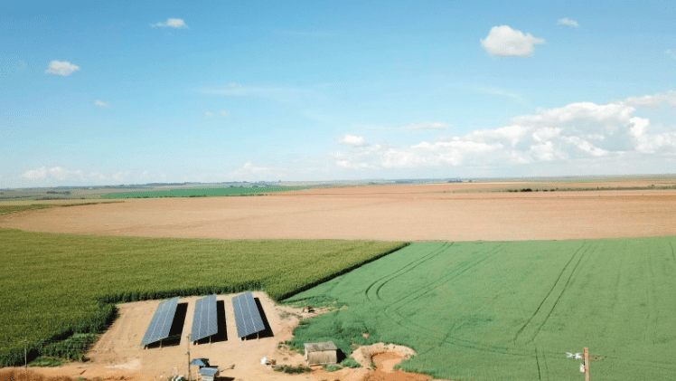 Foto de área de irrigação com pivô central com energia solar. Na foto, três placas grandes de energia solar estão posicionadas no solo, levemente inclinadas.