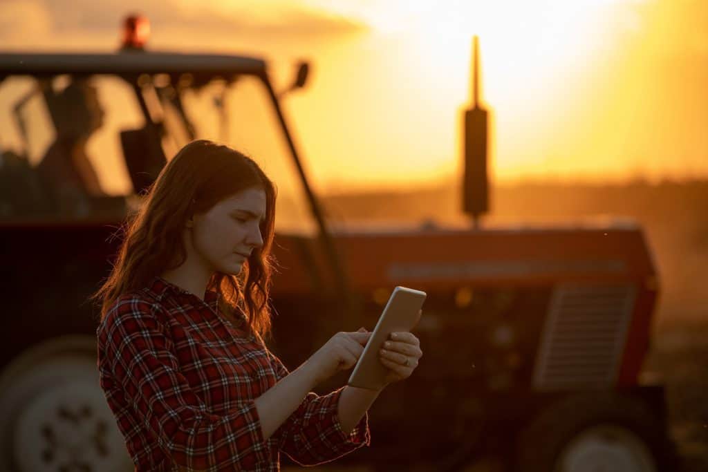 Foto de produtora rural na lavoura, segurando um tablet. Atrás dela há uma máquina e o pôr do sol