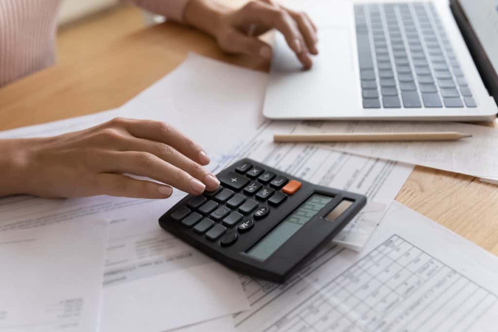 Foto de mãos sobre uma mesa, usando a calculadora em primeiro plano. No segundo plano, há um computador e alguns papéis sobre a mesa.