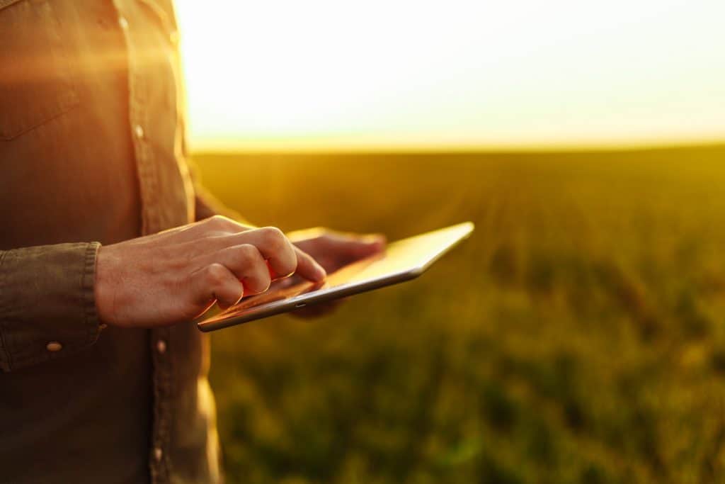 Foto de homem segurando um tablet no meio da lavoura, atrás do pôr-do-sol