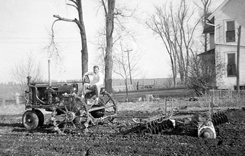Foto em preto e branco de uma carroça com um homem sentado sobre a roda, em uma lavoura
