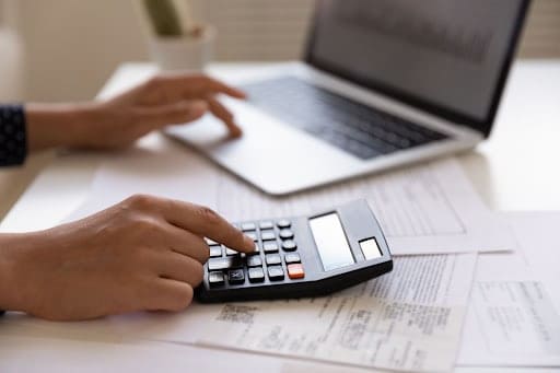 BPO financeiro: foto de duas mãos sobre uma mesa de trabalho. Uma mão calcula em uma calculadora, a outra digita algo em um notebook. Sobre a mesa, há papeis espalhados.