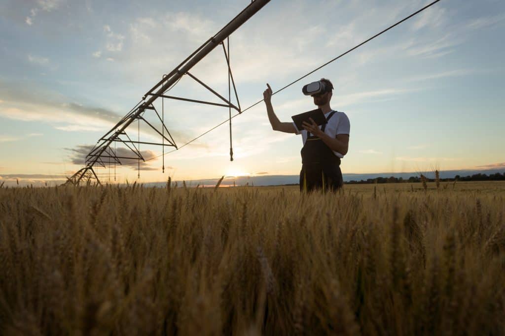 Foto de agricultor na lavoura, com óculos de realidade virtual e tablet. Ele está em baixo de um sistema de irrigação central.