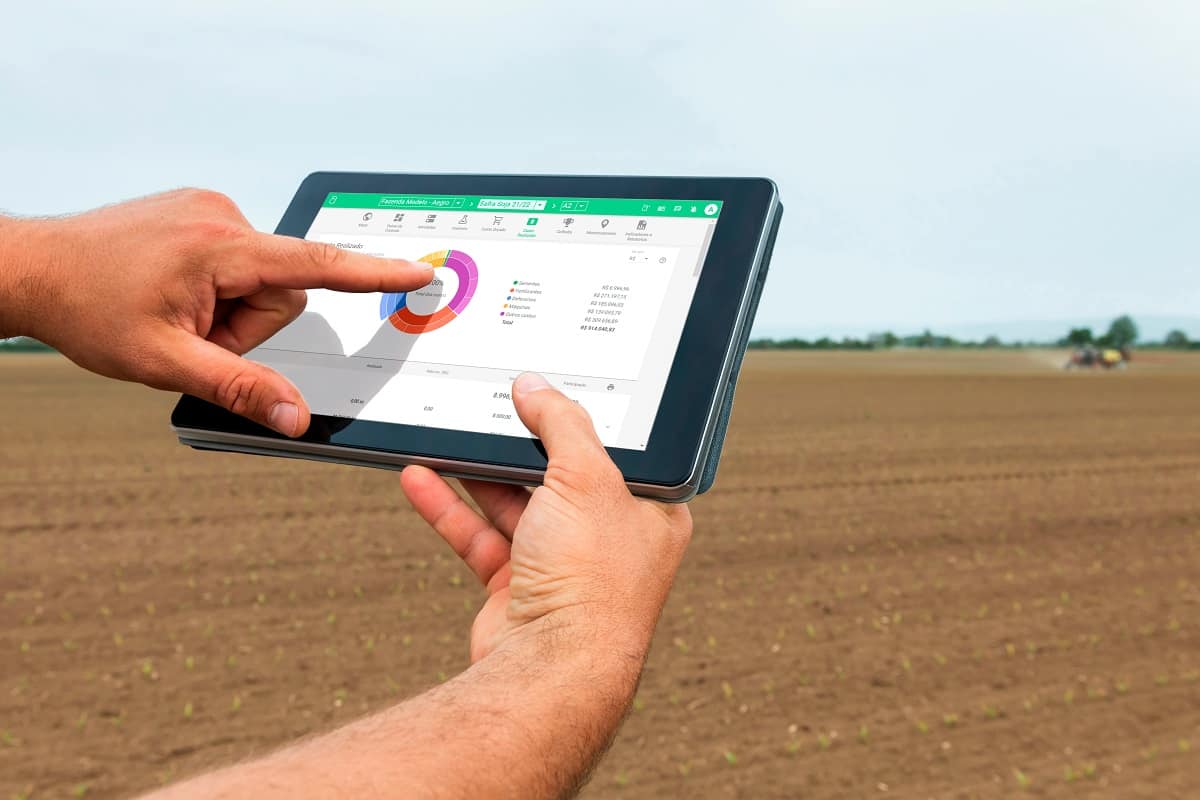 controle financeiro da fazenda: foto de mãos de homem mexendo em um tablet, com uma fazenda no fundo