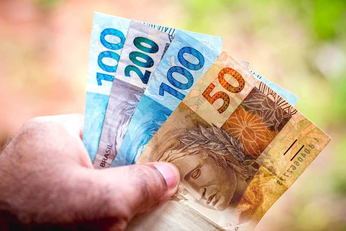 Foto de mão de um senhor segurando notas de 100, 200 e 50 reais, com uma fazenda no fundo