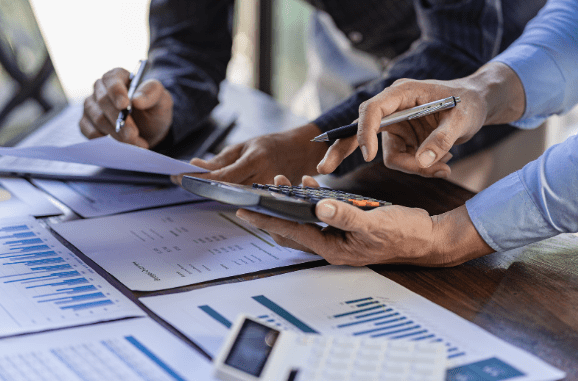 Duas pessoas analisando documentos financeiros com gráficos e tabelas, utilizando uma calculadora e fazendo anotações com canetas. O ambiente sugere um escritório ou local de trabalho administrativo