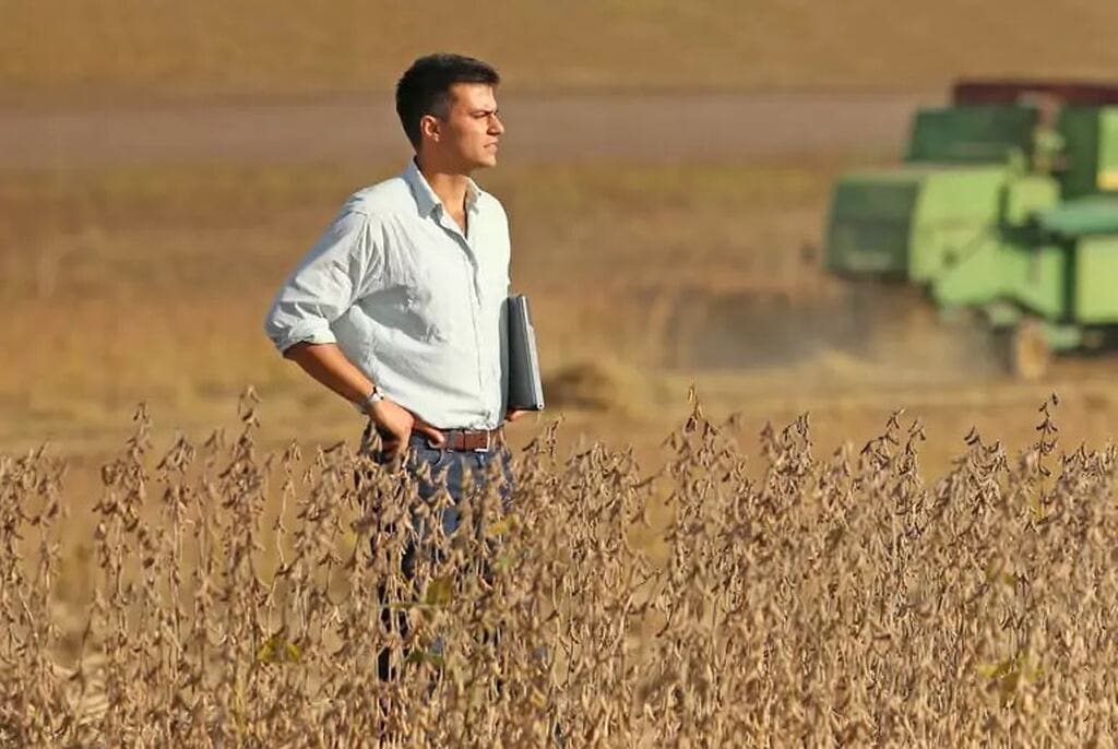 Produtor rural em pé em meio à plantação de soja, segurando um notebook e observando colheitadeiras trabalhando ao fundo.