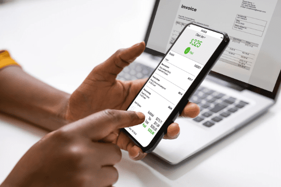 Pessoa segurando um smartphone com a tela exibindo uma nota fiscal eletrônica. Ao fundo, um notebook aberto mostra a mesma fatura digitalizada na tela.